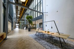 Moderne kantoorlobby aan de H.J.E. Wenckebachweg met hoge plafonds, glazen gevel, en een lange houten vergadertafel onder hanglampen.